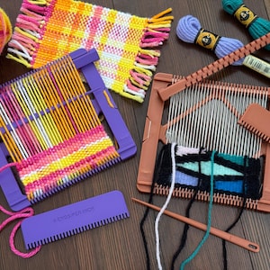 May include: A selection of weaving tools and yarn in various colours. Includes weaving looms, yarn, and combs. The woven projects display vibrant hues of pink, yellow, orange, and blue. The tools are arranged on a wooden surface.