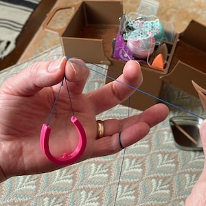 May include: A pink, horseshoe-shaped tatting shuttle held in a hand, with blue thread looped around the fingers. A wooden shuttle with blue thread is in the other hand. A brown craft box is in the background.