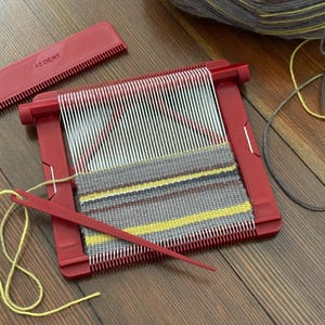Könnte beinhalten: Roter Webrahmen mit weißen und roten Fäden, teilweise mit grauem, gelbem und braunem Garn gewebt. Ein roter Kamm und eine Nadel sind enthalten. Ein Knäuel Garn befindet sich im Hintergrund. Der Webstuhl steht auf einer Holzoberfläche.