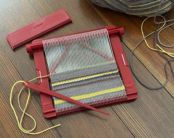 Peut inclure: Métier à tisser rouge avec des fils blancs et rouges, partiellement tissé avec du fil gris, jaune et marron. Un peigne et une aiguille rouges sont inclus. Une pelote de laine est en arrière-plan. Le métier est sur une surface en bois.