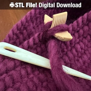 May include: A close-up of a burgundy knitted fabric with a white bone knitting needle and a wooden stitch marker. The stitch marker is shaped like a fish and has a comb-like design on the top.