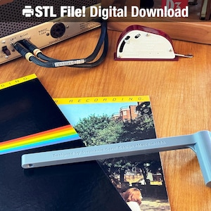 May include: A gray 3D printed tool with the text "Designed by Stephanie Gervi Design - Made in USA" on it. The tool is lying on top of two vinyl records, one with a rainbow stripe and the other with a picture of a building.