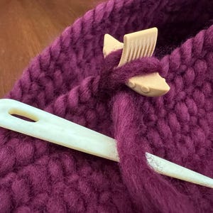 May include: A close-up of a burgundy knitted fabric with a wooden stitch marker and a white bone knitting needle. The stitch marker is shaped like a fish and has a row of teeth on the top.
