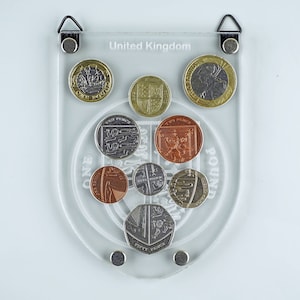 Puede incluir: Un expositor de monedas de acrílico transparente con fondo blanco. El expositor presenta un recorte en forma de escudo con las palabras "Reino Unido" y "Una Libra" impresas en él. El expositor contiene una colección de monedas británicas, incluyendo una moneda de 50 peniques, una moneda de dos peniques, una moneda de un penique y una moneda de dos libras.