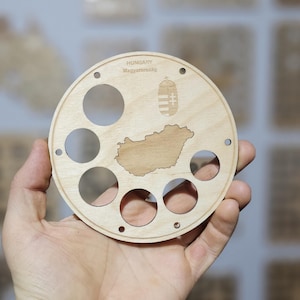 May include: A round wooden item with cut-out circles and a map of Hungary. The text "HUNGARY Magyarország" is engraved at the top, along with a coat of arms. The item is held in a hand.