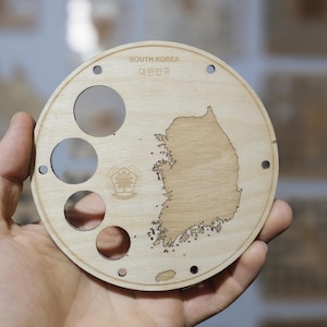 May include: A circular wooden plaque with a laser-cut map of South Korea. The plaque has three circular cutouts and the text "SOUTH KOREA" and "대한민국". Several small holes are around the edge of the plaque.