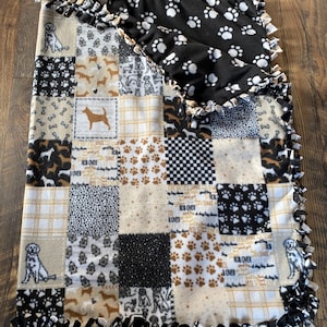 May include: A patchwork fleece blanket featuring various dog-themed patterns. The blanket showcases squares with dog silhouettes, paw prints, and bone designs in shades of brown, black, and cream. The edges are finished with a knotted fringe.