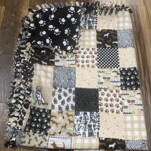 May include: A patchwork fleece blanket featuring various dog-themed patterns. The blanket includes squares with paw prints, dog silhouettes, and the words "Roll Over". The edges are finished with knotted fringe.
