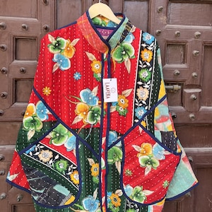 Puede incluir: Una chaqueta acolchada con un diseño de patchwork vibrante, con paneles rojos y verdes adornados con coloridos estampados florales. La prenda tiene cuello mao, mangas largas, dos bolsillos delanteros grandes y ribetes azul oscuro. Una etiqueta blanca dice LAAVEKA.