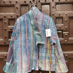 May include: A blue jacket featuring a multi-coloured striped pattern with intricate kantha stitching throughout. The garment has a wide collar, long sleeves, and yellow tie closures on the side. A white tag with the text LAAVEKA is attached.