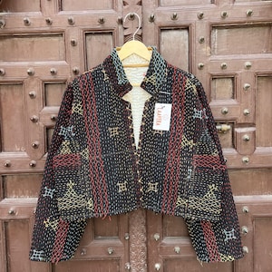 Op de afbeelding: Een zwart doorgestikt jack met ingewikkeld kantha-borduurwerk in rode, gele en blauwe geometrische patronen. Het jack heeft lange mouwen, een opstaande kraag en een witte voering in sherpa-stijl. Een wit label met de tekst LAAVERA is bevestigd.