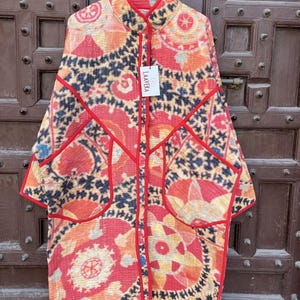 May include: A long-sleeved quilted jacket featuring a vibrant pattern of red, orange, and black circular motifs. The garment has a high collar, two large front pockets, and bright red piping along the edges. A white tag labeled LAAVEKA is attached.