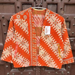May include: An orange, open-front jacket with a unique patchwork design. The jacket features a variety of patterns, including zigzags, floral motifs, and geometric shapes. The interior lining has a zebra-like pattern. A tag with the text "LAAYEKA" is attached.