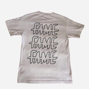 May include: A light beige t-shirt with the word "FAME" printed three times in black, stacked vertically on the back. The design features a stylized, angular font. The shirt has short sleeves and a crew neck.