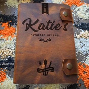May include: A brown leather recipe book with the name "Katie's" and "Favorite Recipes" engraved on the cover. The book has a strap closure and the text "Home Cooking" with a fork, spoon, and knife design. The color is described as "old whiskey color".