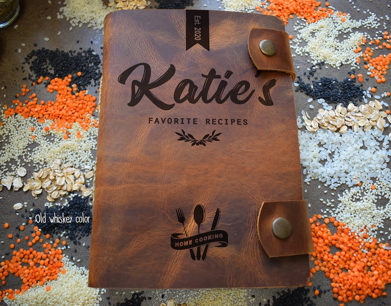 May include: A brown leather recipe book with the name "Katie's" and "Favorite Recipes" engraved on the cover. The book has a strap closure and the text "Home Cooking" with a fork, spoon, and knife design. The color is described as "old whiskey color".
