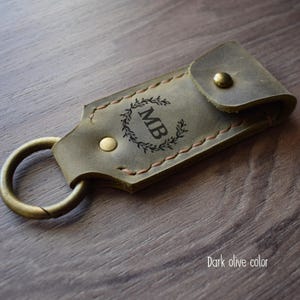 Può includere: Un portachiavi in ​​cuoio verde oliva scuro con un anello per le chiavi e una chiusura a lembo. Il portachiavi è personalizzato con le iniziali "MB" in un design a corona.