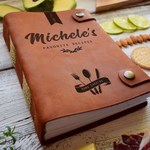 May include: A brown leather recipe book with the text "Michele's Favorite Recipes" and a "Home Cooking" graphic. The book has a button closure and is surrounded by food items like avocado, tomatoes, and crackers.