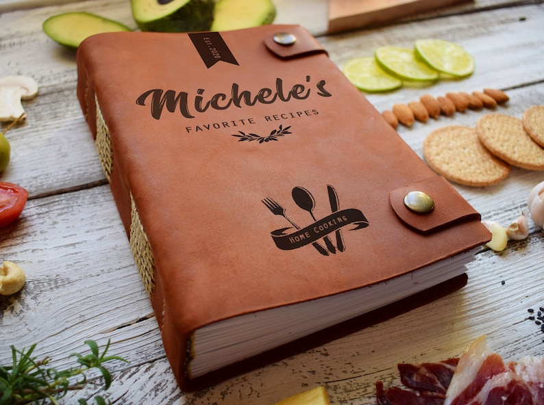 May include: A brown leather recipe book with the text "Michele's Favorite Recipes" and a "Home Cooking" graphic. The book has a button closure and is surrounded by food items like avocado, tomatoes, and crackers.