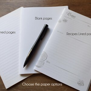 May include: Three white paper booklets with different interior layouts: one with plain lined pages, one with blank pages, and one with a printed recipe template featuring sections for ingredients, method, and notes. A black ballpoint pen rests on top.