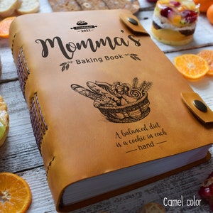 May include: Camel-colored leather-bound cookbook titled "Momma's Baking Book" with a basket of bread illustration. The book has a snap closure and stitched spine. The text reads "A balanced diet is a cookie in each hand."