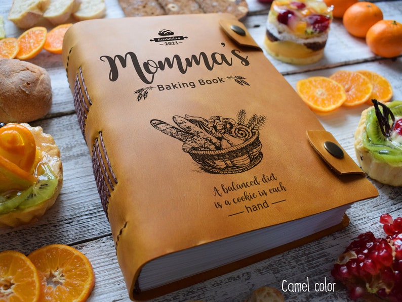 May include: Camel-colored leather-bound cookbook titled "Momma's Baking Book" with a basket of bread illustration. The book has a snap closure and stitched spine. The text reads "A balanced diet is a cookie in each hand."