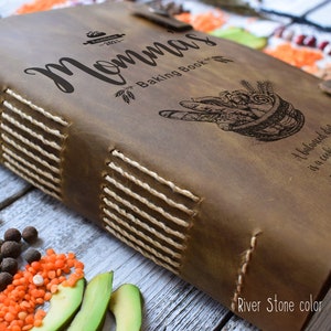 May include: A brown leather-bound "Momma's Baking Book" with stitched binding and a clasp closure. The cover features engraved text and illustrations, including the phrase "A balanced diet is a cookie in each hand."