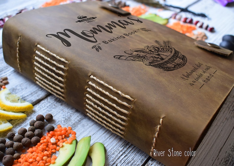 May include: A brown leather-bound "Momma's Baking Book" with stitched binding and a clasp closure. The cover features engraved text and illustrations, including the phrase "A balanced diet is a cookie in each hand."