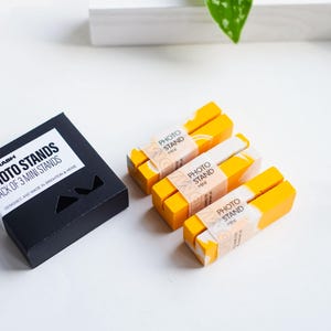 May include: A black box with the text "PHOTO STANDS PACK OF 3 MINI STANDS" is next to three yellow and white photo stands. Each stand has a label saying "PHOTO STAND HERE". These stands are designed for photography.