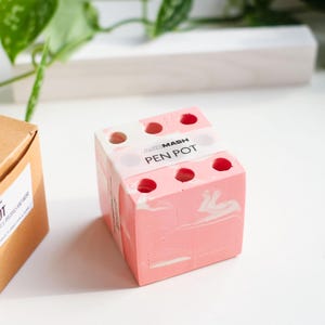 May include: A pink and white marbled pen pot with the text "PEN POT" on a white label. The pen pot has six holes for pens and pencils. A brown box with a label is to the left of the pen pot.