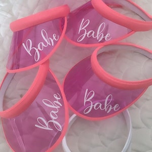 May include: A collection of clear, pink visors with a coral-colored trim. Each visor is printed with the word "Babe" in a white script font. One white visor is printed with the word "Bride".