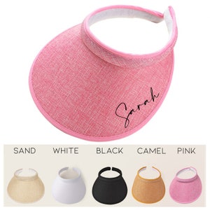 May include: Pink sun visor with a black embroidered name "Sarah" on the front. The visor is made of a woven fabric and has a black elastic band.