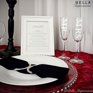 May include: Two champagne flutes with the words "MR." and "MRS." etched on them. The flutes are on a red table cloth with a clear glass charger plate. A white plate with a black napkin is in the foreground. A white framed paper with a list of names is in the background.
