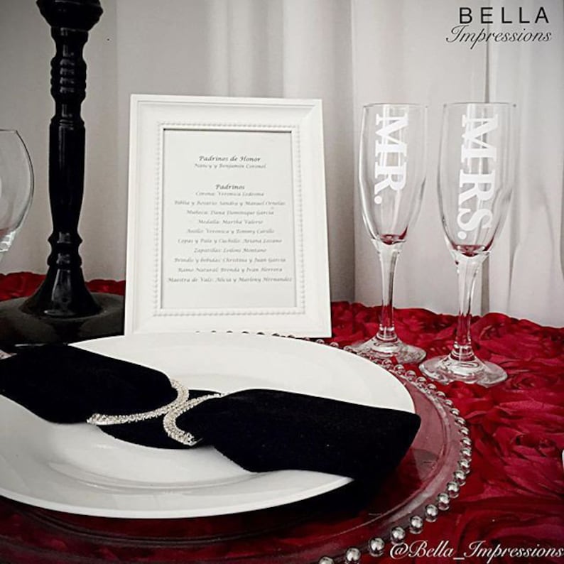 May include: Two champagne flutes with the words "MR." and "MRS." etched on them. The flutes are on a red table cloth with a clear glass charger plate. A white plate with a black napkin is in the foreground. A white framed paper with a list of names is in the background.