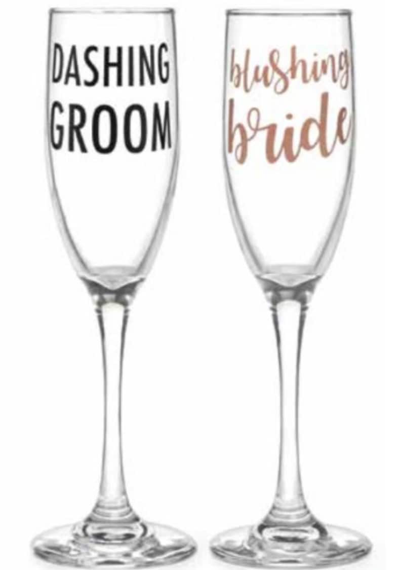 May include: Two champagne flutes with the words "Dashing Groom" and "Blushing Bride" in black and rose gold lettering.