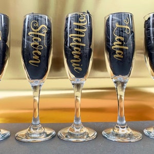 May include: Five champagne flutes with gold lettering on the front. The names on the flutes are: Zoe, Steven, Melanie, Leila, and Carol.