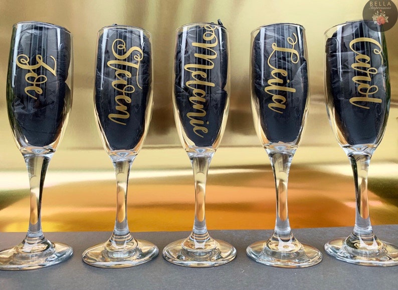 May include: Five champagne flutes with gold lettering on the front. The names on the flutes are: Zoe, Steven, Melanie, Leila, and Carol.