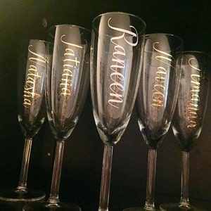 May include: Five clear glass champagne flutes with gold lettering. The names Natasha, Jatitia, Raneen, Maureen, and Tamara are written on the flutes.