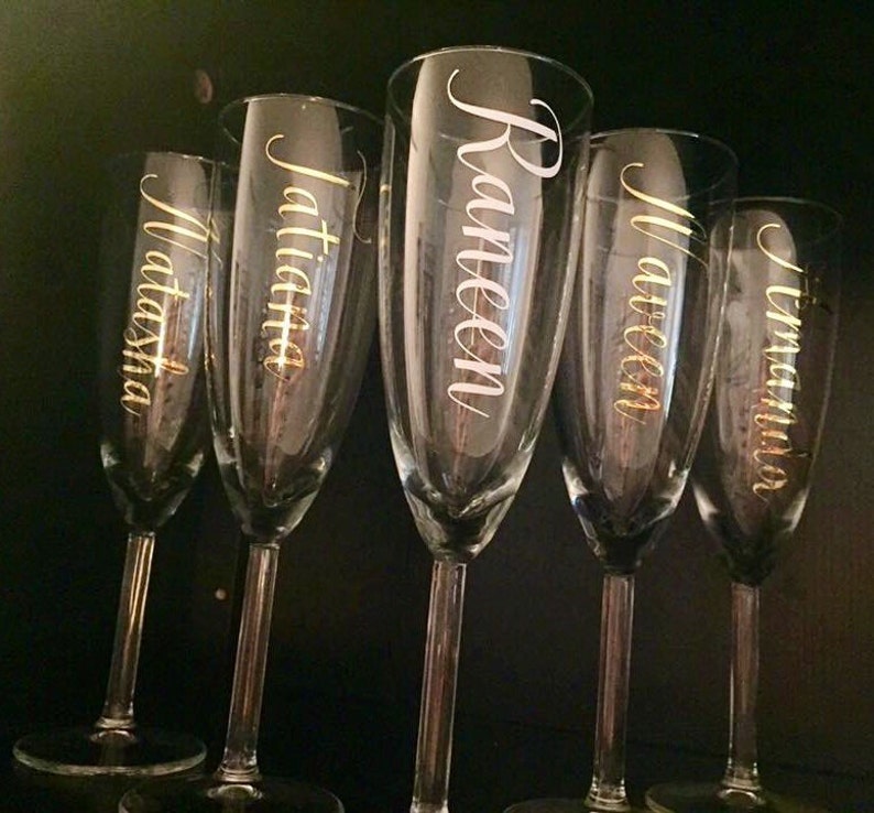 May include: Five clear glass champagne flutes with gold lettering. The names Natasha, Jatitia, Raneen, Maureen, and Tamara are written on the flutes.