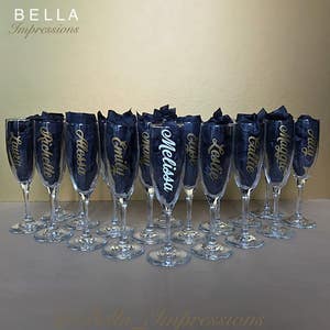 May include: A set of 14 clear glass champagne flutes with gold lettering on each glass. The names on the glasses are Laura, Richelle, Alessa, Emily, mmMo, Melissa, Gyoe, Leslie, Callie, Maggie, Haly.