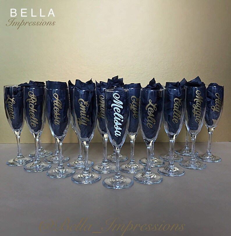 May include: A set of 14 clear glass champagne flutes with gold lettering on each glass. The names on the glasses are Laura, Richelle, Alessa, Emily, mmMo, Melissa, Gyoe, Leslie, Callie, Maggie, Haly.