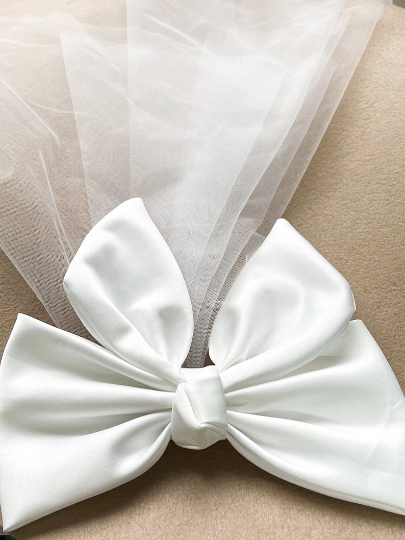 May include: A white satin bow with a tulle veil attached. The bow is tied in a knot and the tulle is draped over the bow.