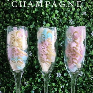May include: Three clear glass champagne flutes filled with pink and blue cotton candy. Each flute has a gold sticker with the text "Olivia 07.06.19".