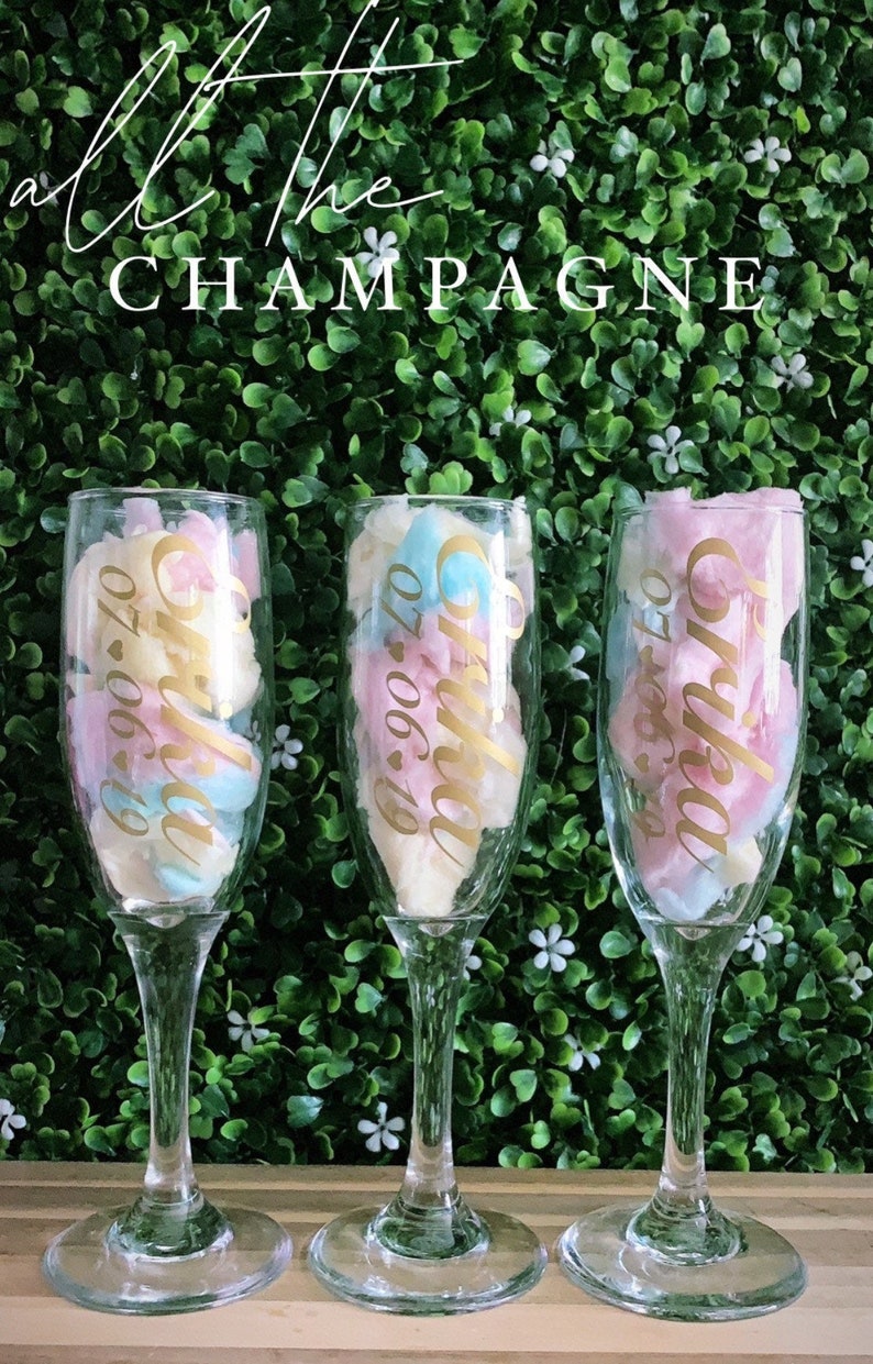 May include: Three clear glass champagne flutes filled with pink and blue cotton candy. Each flute has a gold sticker with the text "Olivia 07.06.19".