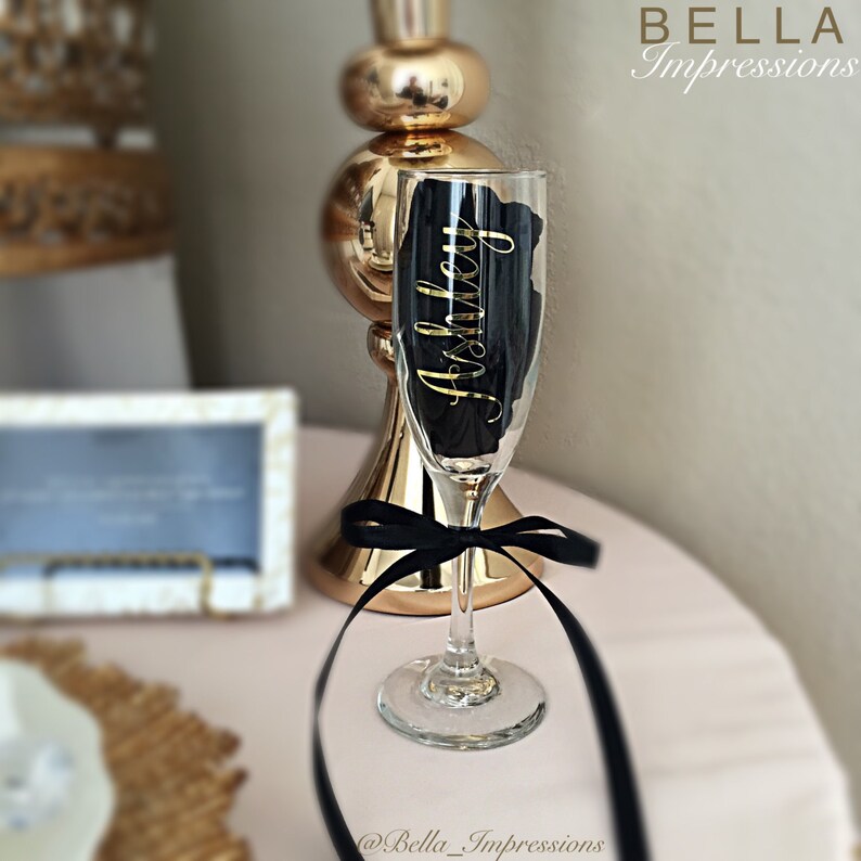 May include: A champagne flute with a black and gold personalized label that says "Ashley". The flute is tied with a black ribbon and is sitting on a pink surface.