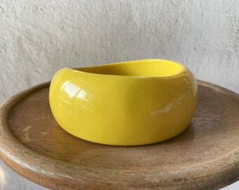 May include: A vibrant yellow, rounded bangle bracelet. The bracelet is made of a glossy material and rests on a wooden, round tray. The bracelet's smooth surface reflects light, highlighting its vibrant colour and simple design. The bracelet is a bold fashion accessory.