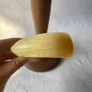 Pode incluir: Uma pulseira de resina amarela clara e marmorizada. A pulseira tem uma forma arredondada e lisa e um acabamento brilhante. A cor é um amarelo quente e cremoso com padrões em espiral. A pulseira é segurada em frente a um objeto de madeira.