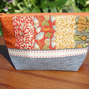 Può includere: Una pochette con cerniera in tessuto patchwork con fondo in denim grigio e un nastro bianco con cuciture rosse. La pochette presenta un motivo floreale in tonalità arancio, rosso e giallo.