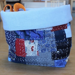 May include: A blue and red patchwork fabric storage bin with a light blue fabric top. The bin is filled with fabric scraps and a pair of orange scissors.