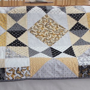 Op de afbeelding: Een quilt met een patchwork-ontwerp met verschillende stoffen in grijstinten, zwart en geel. De quilt is gemaakt van vierkanten en driehoeken, en de stoffenpatronen omvatten vlinders, bijen en geometrische ontwerpen.